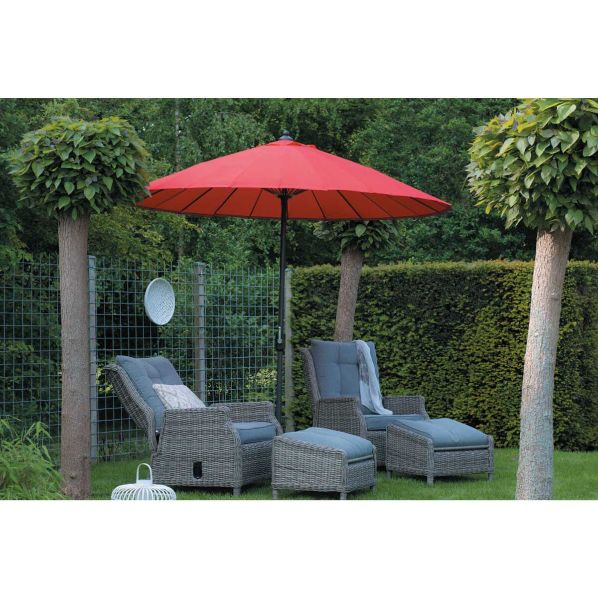 A garden scene with red umbrella, wicker chairs, and trees. Green lawn and fence.