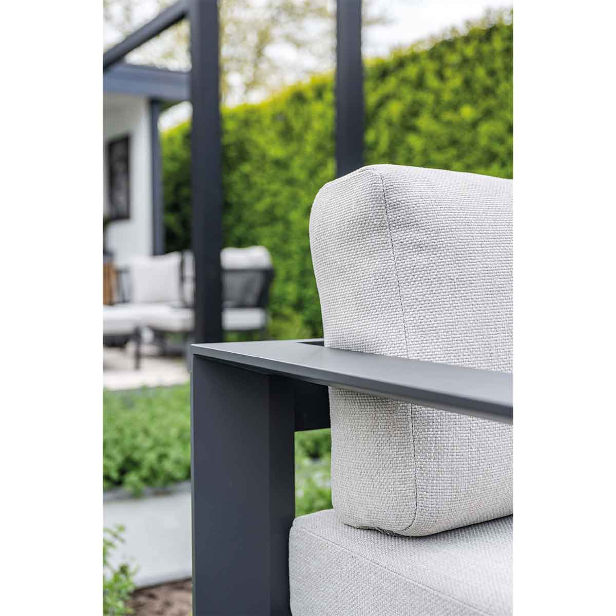 Close-up of a light gray outdoor sofa with dark gray frame. Green hedge in the background.