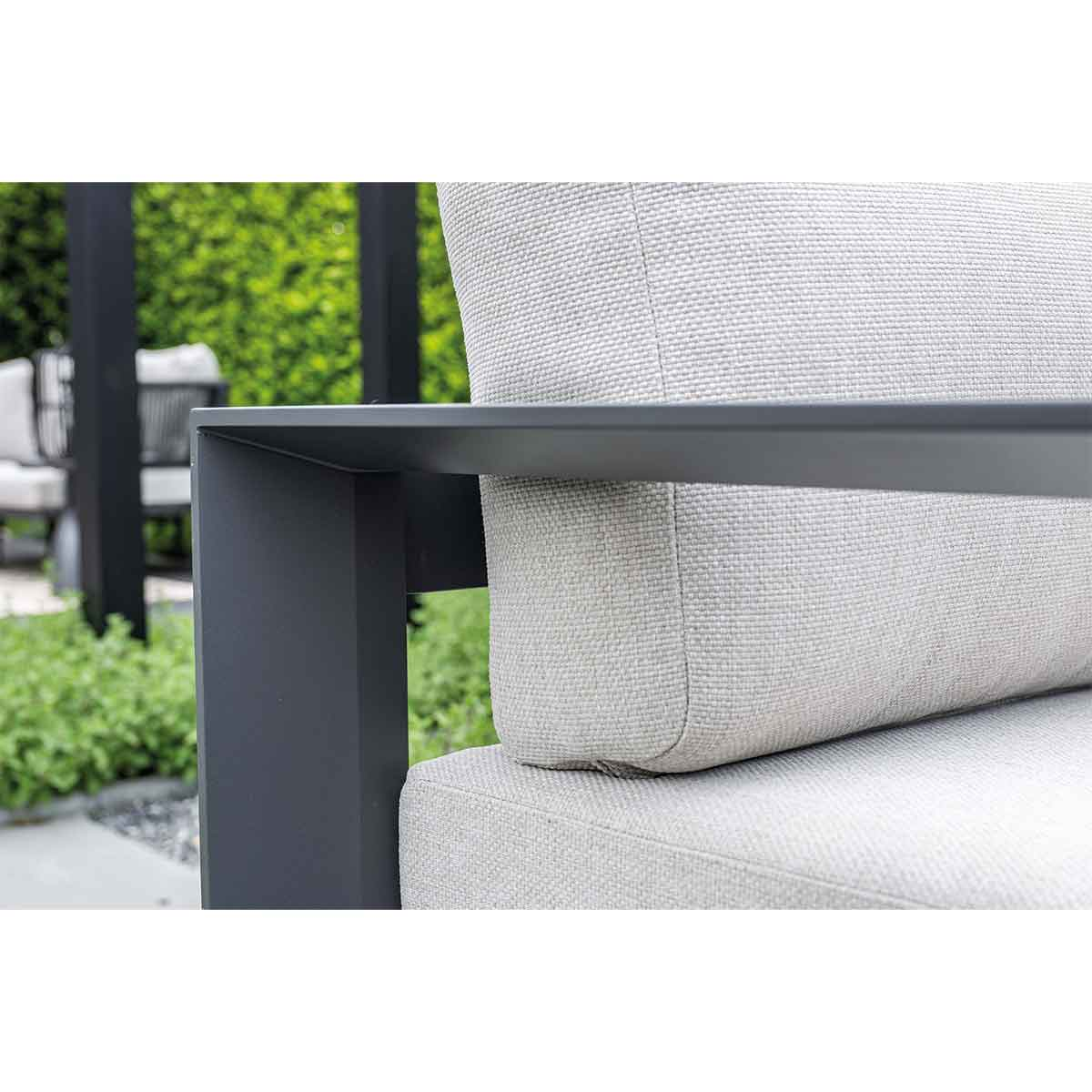 Close-up of outdoor sofa. Light gray cushion, dark gray frame. Green hedge in the background.