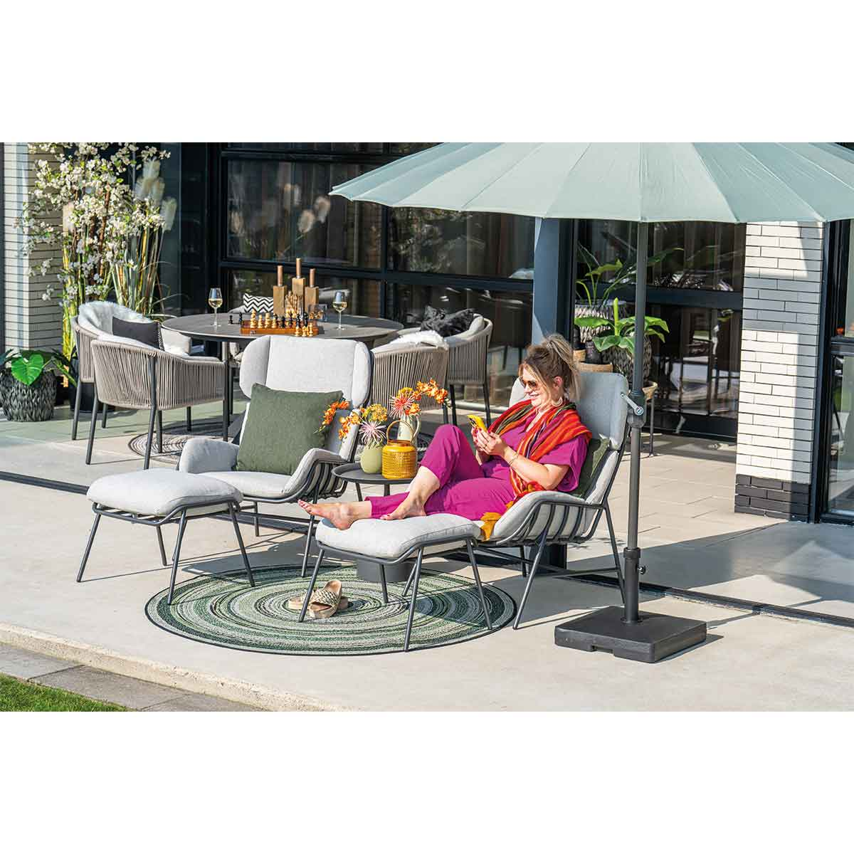 Woman relaxing on outdoor furniture, reading a phone. Table with drinks and snacks. Green rug, plants, and umbrella.