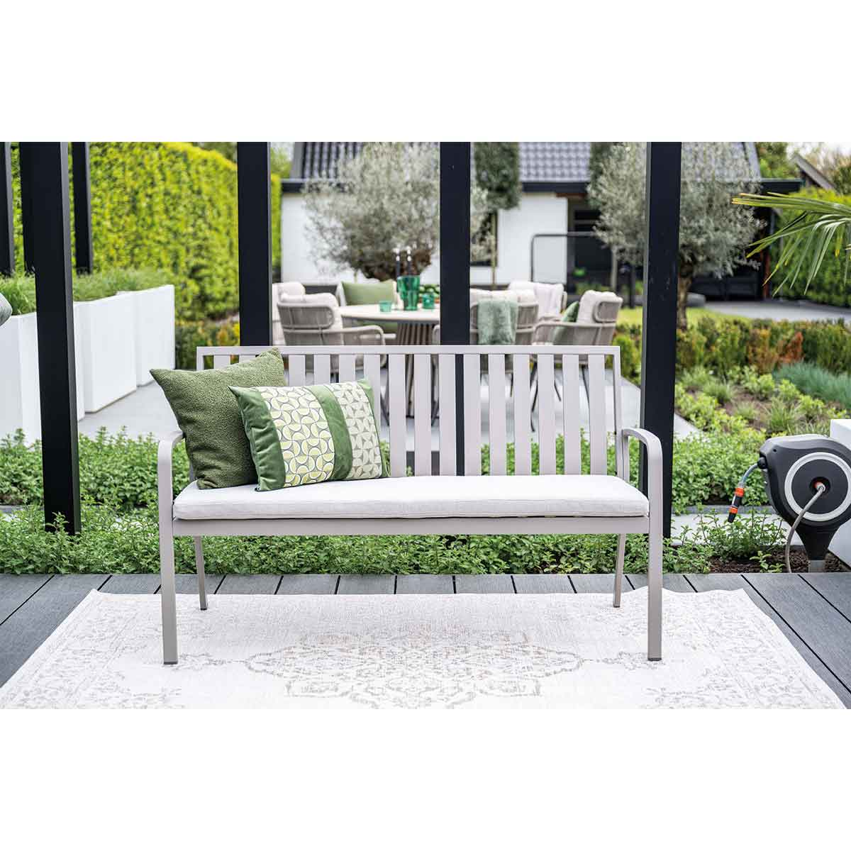 Banc gris avec coussins et oreillers verts, fond de jardin.
