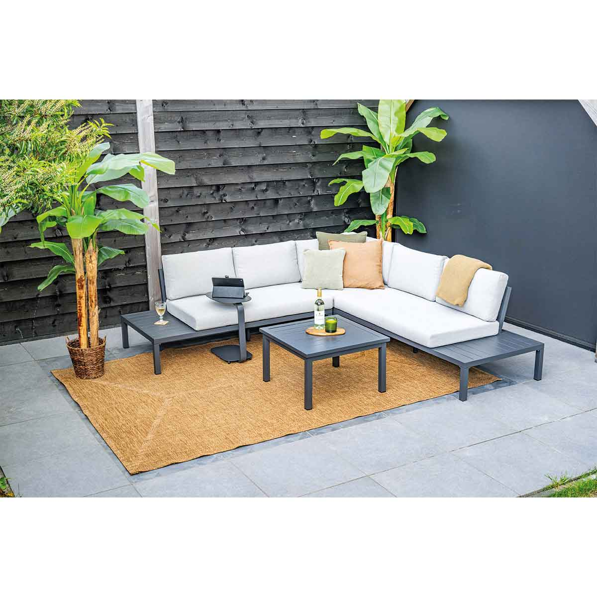 Outdoor sectional sofa with tables, rug, plants. A glass, bottle, and tablet are on the tables.