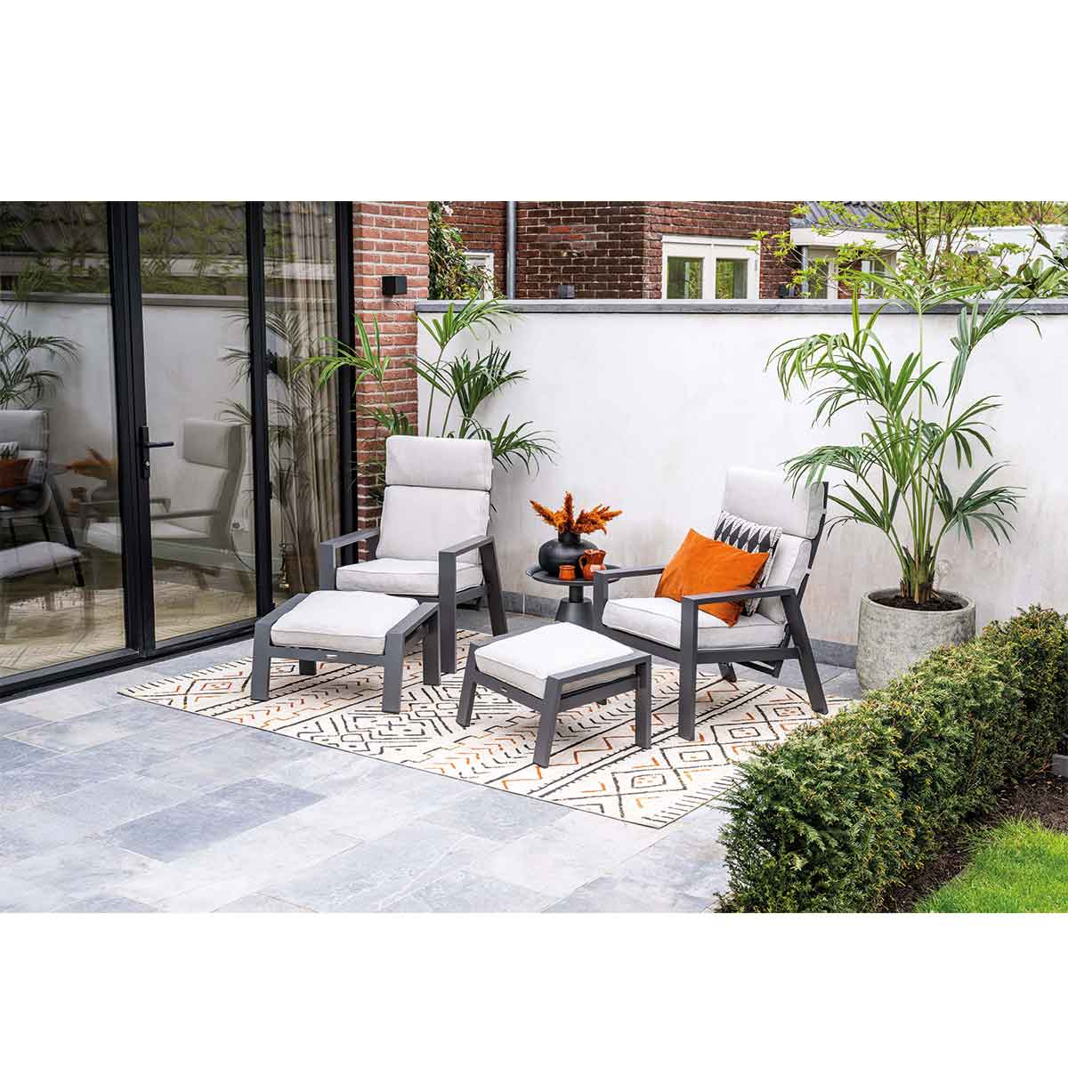 Aménagement de patio extérieur avec deux chaises, des poufs et une table d'appoint sur un tapis à motifs, avec de la verdure.