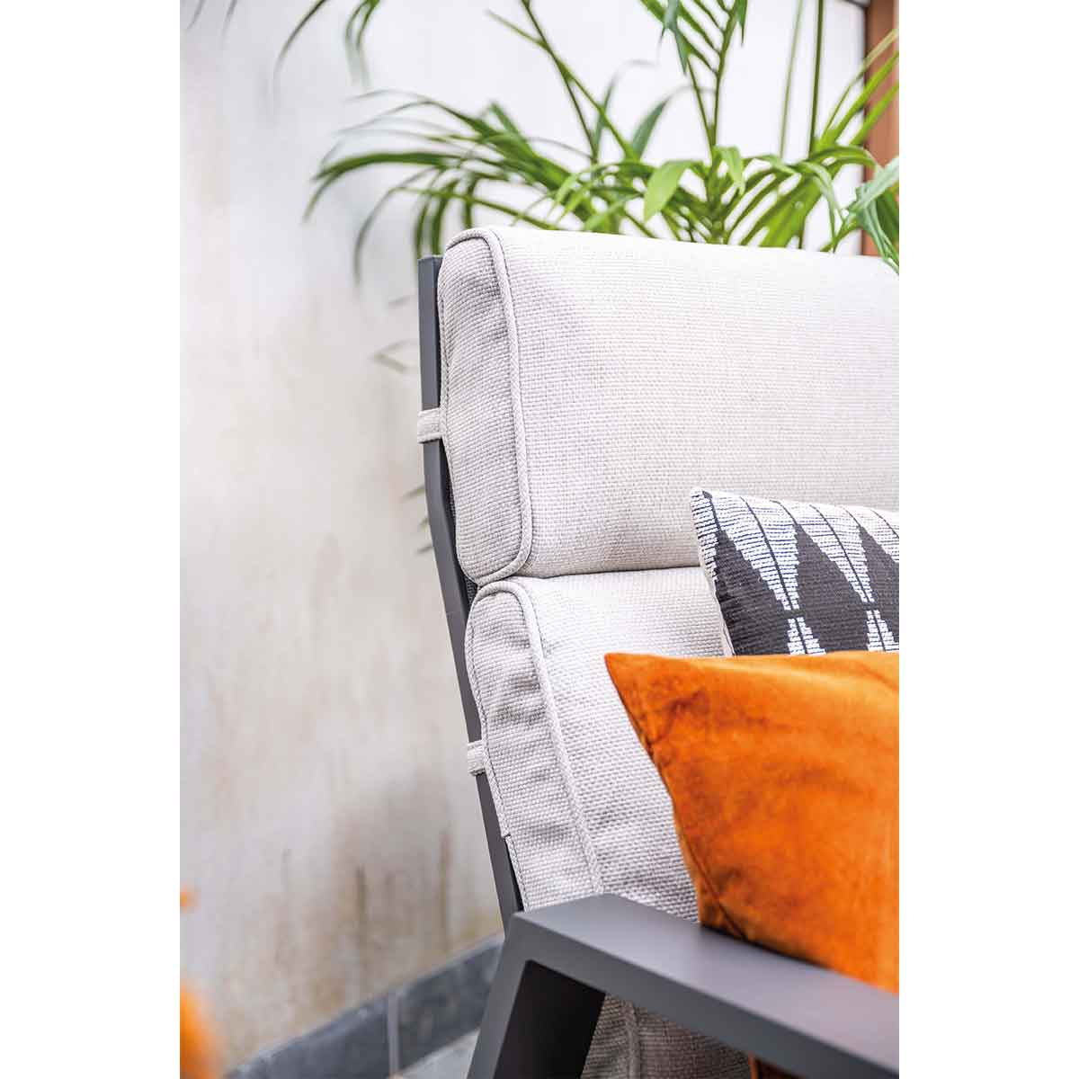 Gros plan sur une chaise beige d'extérieur avec cadre gris, coussins orange et noirs, et plantes vertes en arrière-plan.