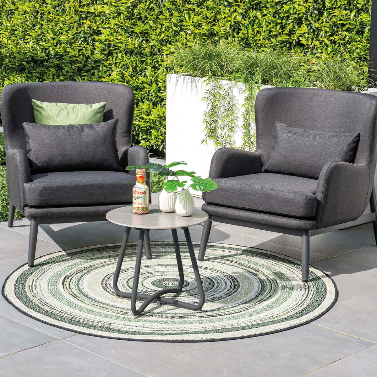 Twee grijze stoelen, een kleine tafel en een kleed op een terras met planten.