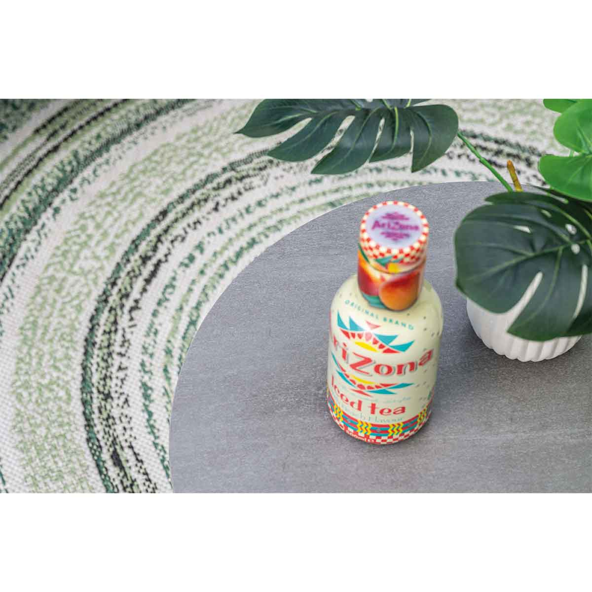 Een Arizona Iced Tea fles op een grijze tafel met een groen-wit tapijt en groene planten eromheen.