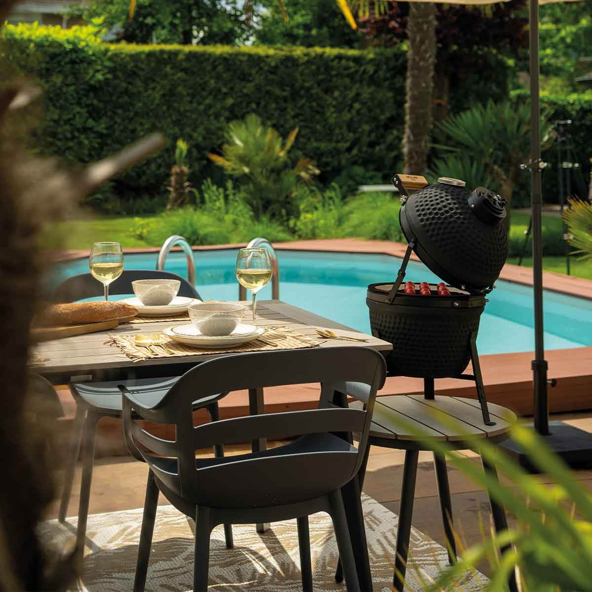 Outdoor dining scene with grill, table set, and a pool in the background.