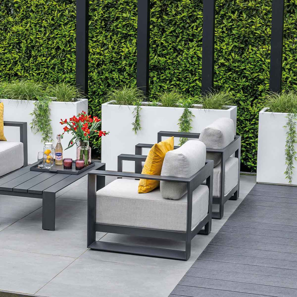 Outdoor seating area with chairs, table, and plants. Green hedge in background.
