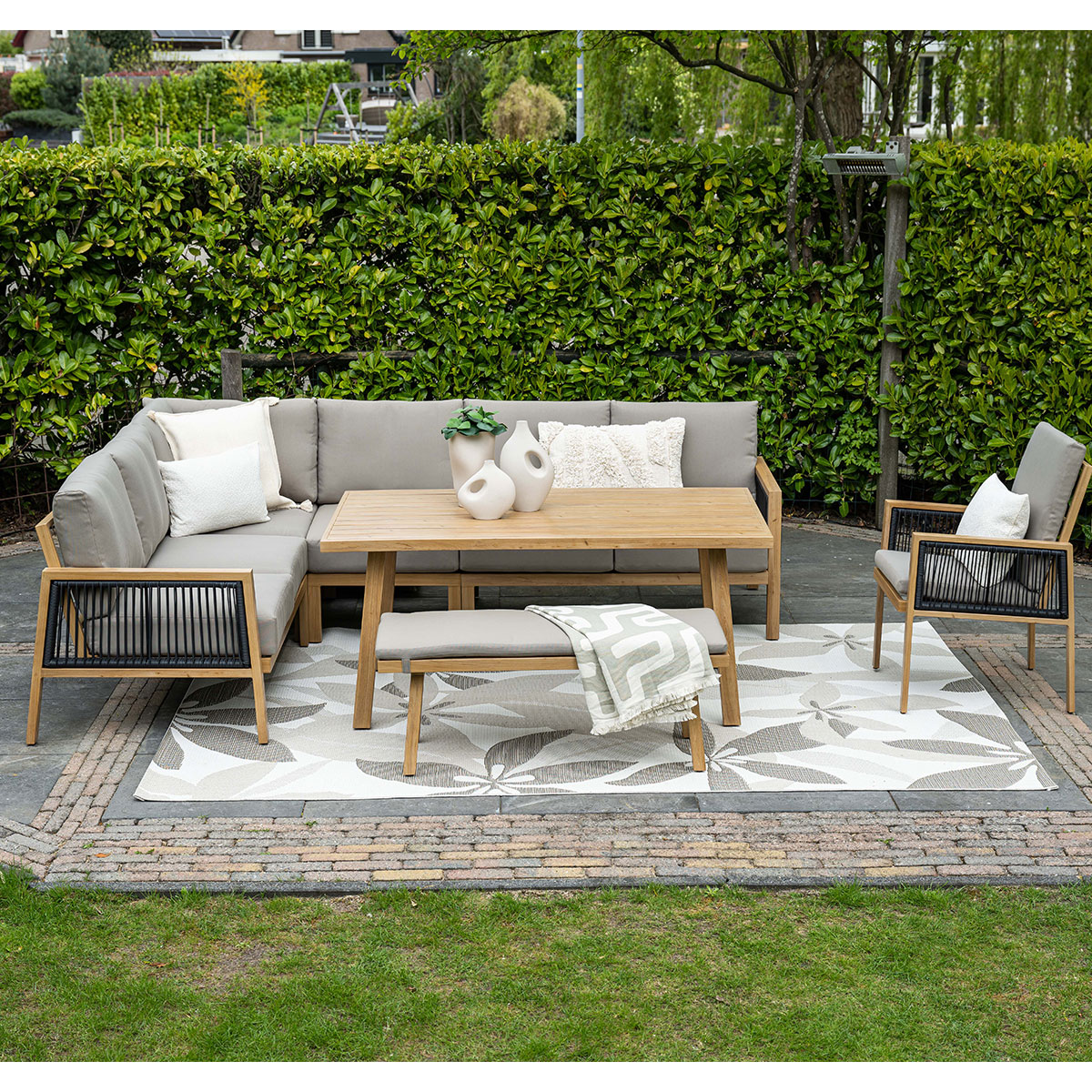 Buitenterrasset: bank, stoelen, tafel en bank. Groene heg achtergrond, bakstenen terras en bloemenkleed.
