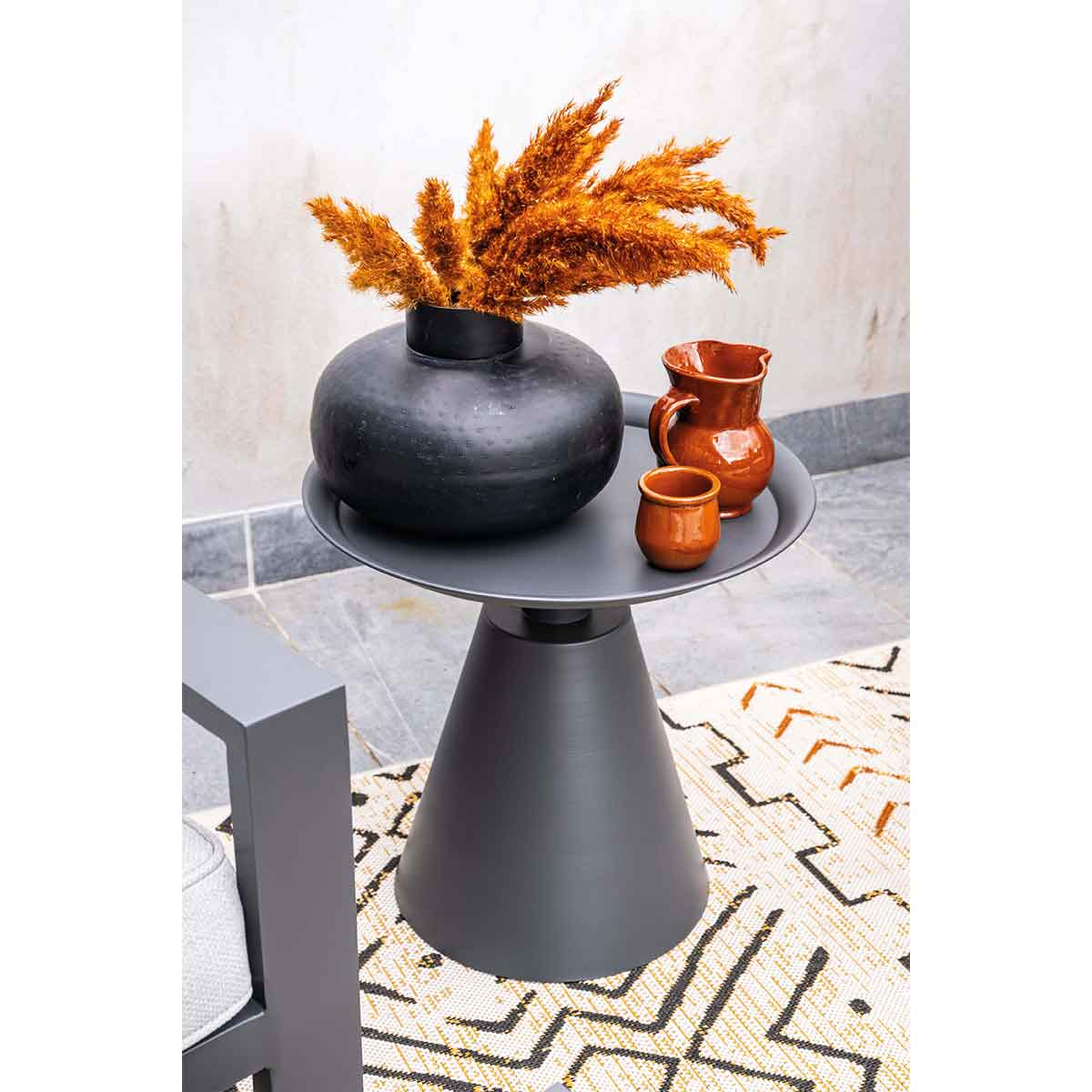 Une table à base noire porte un vase de roseaux orange et deux cruches marron.