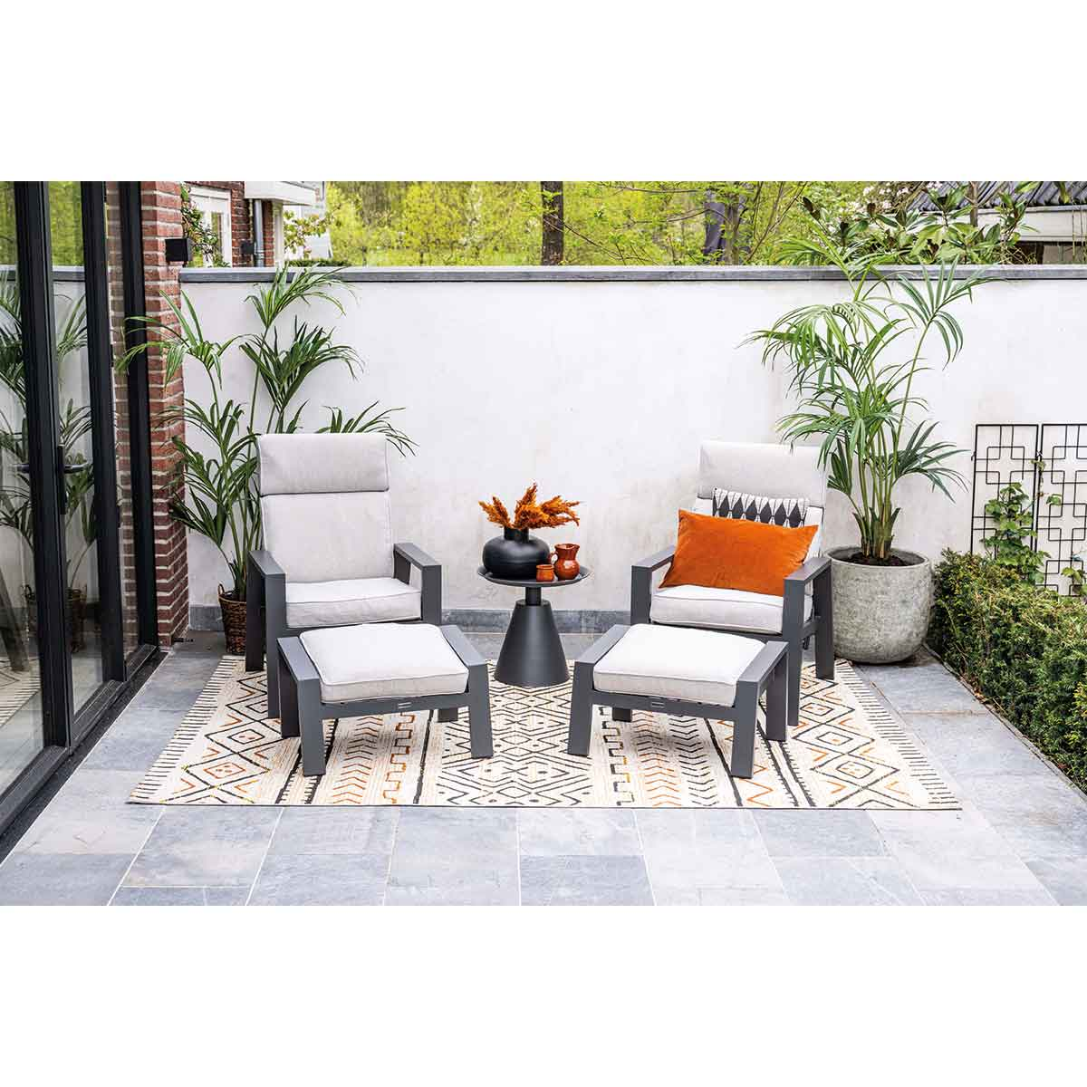Aménagement de patio extérieur avec chaises, tables et plantes. Mobilier gris, tapis à motifs et fond vert luxuriant.