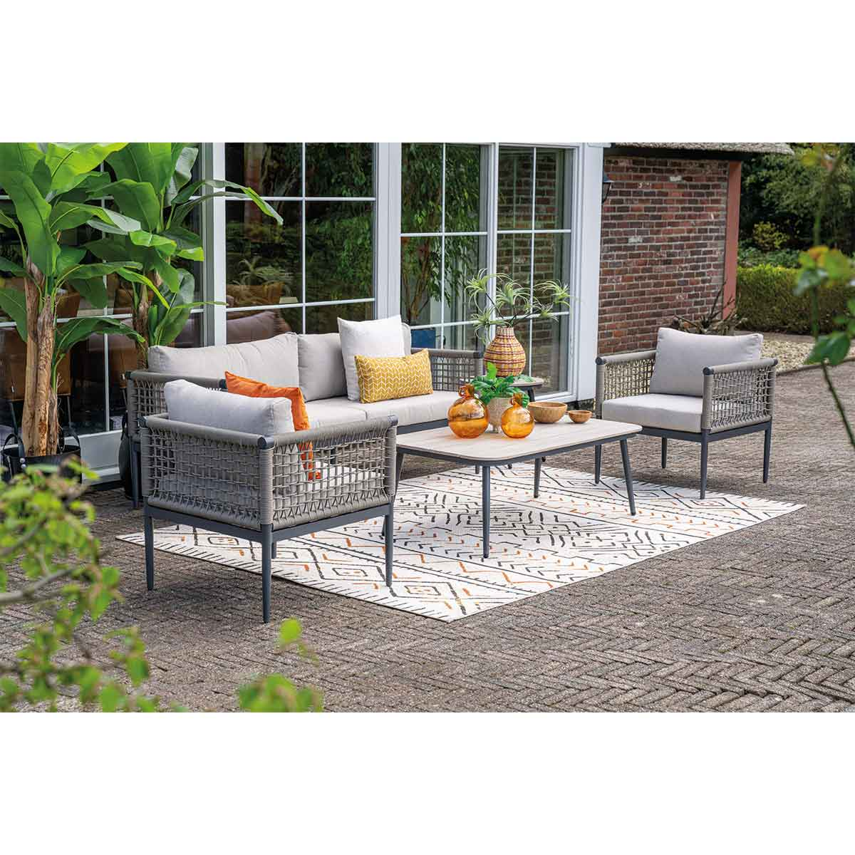 Outdoor patio furniture set. Gray sofa, chairs, table, and rug. People are sitting. Green plants in the background.