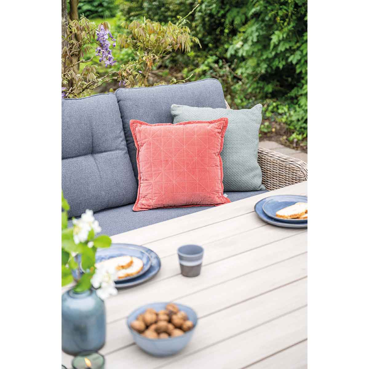 Outdoor table with cushions, food, and flowers. Green foliage in the background.