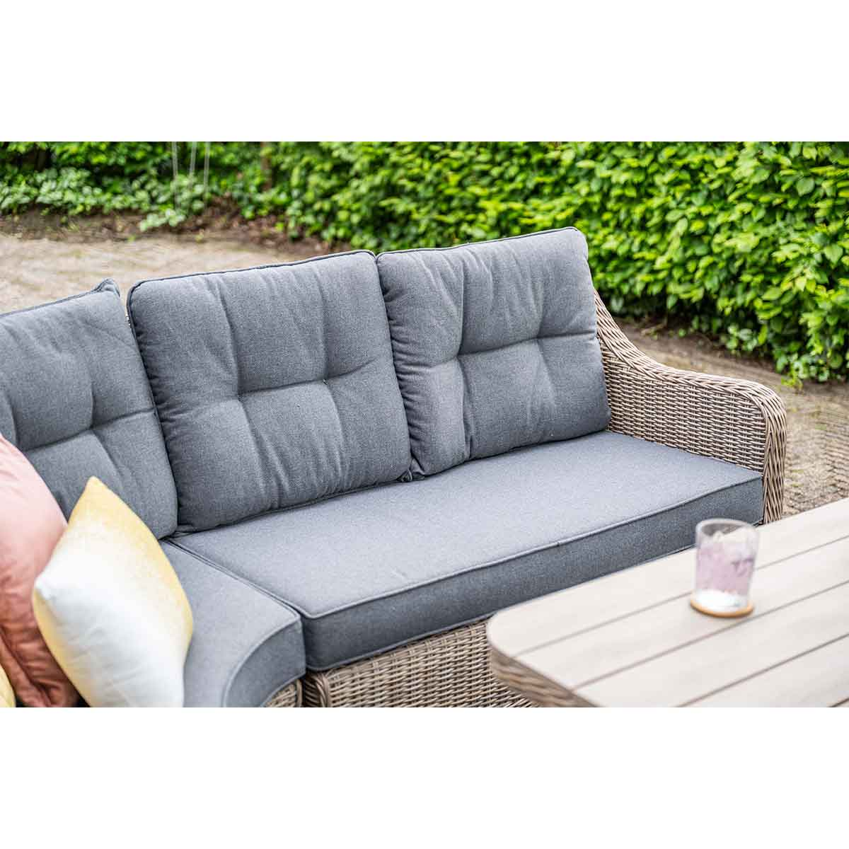 Grey outdoor sofa with cushions, wicker frame. A table with a glass and greenery in the background.