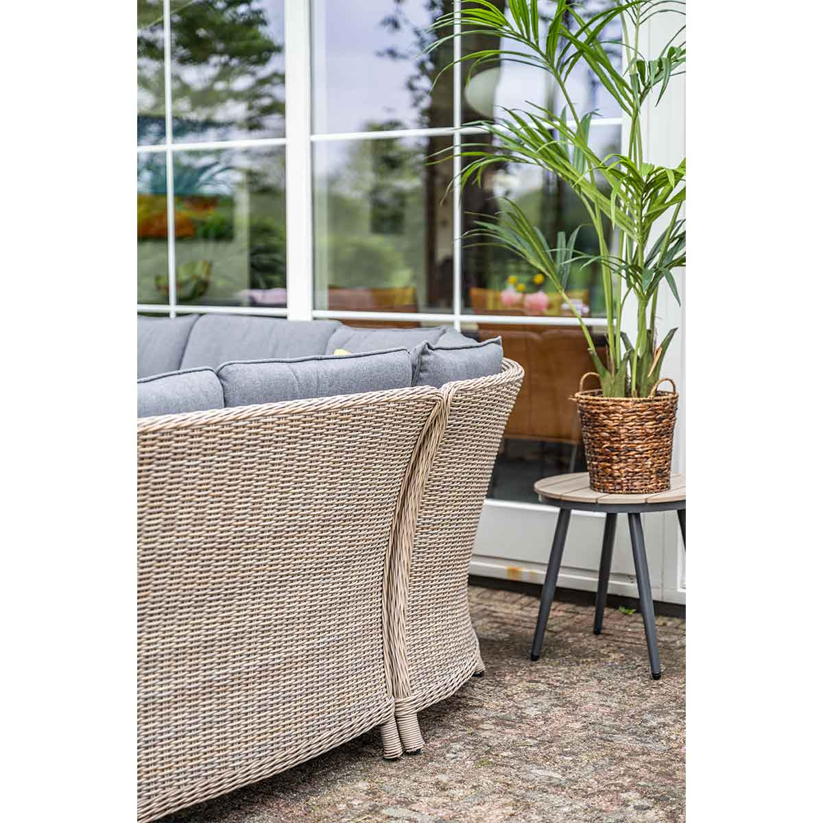 Wicker sofa with grey cushions, table with a plant. Background shows glass windows and greenery.