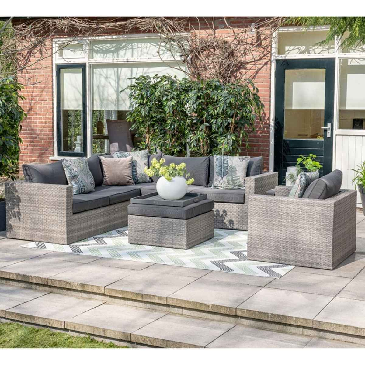 Coin salon extérieur avec meubles en osier gris, tapis et mur de briques en arrière-plan.