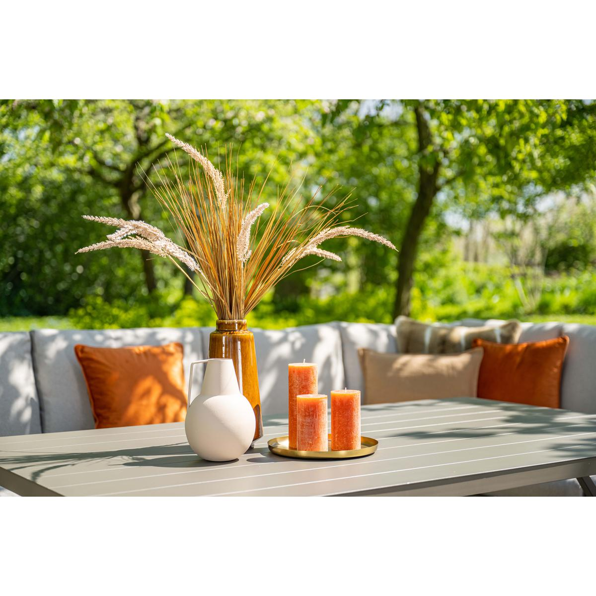 Close-up van kaarsen, vaas en decoratief gras op een grijze tafel, met tuin op de achtergrond.