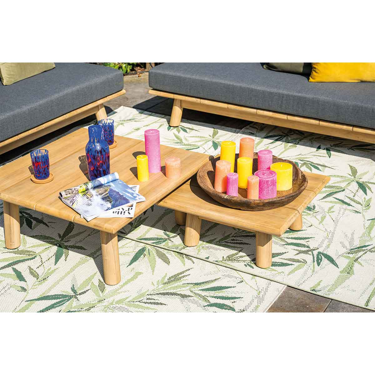 Cadre extérieur avec deux tables en bois, des bougies et des coussins. Tapis à motifs vert et blanc.