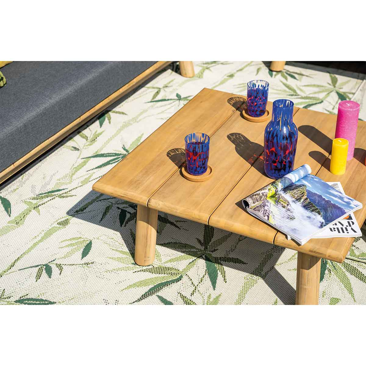Table en bois avec boissons, magazine, bougies et tapis. Bouteilles bleues, feuilles vertes.