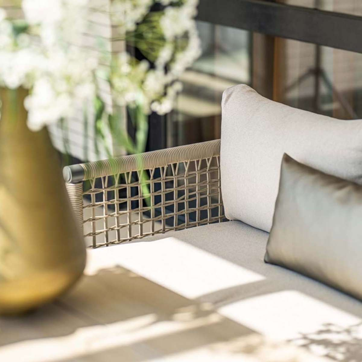 Gros plan sur un mobilier d'extérieur : un canapé avec des coussins clairs et un vase avec des fleurs. Soleil brillant.
