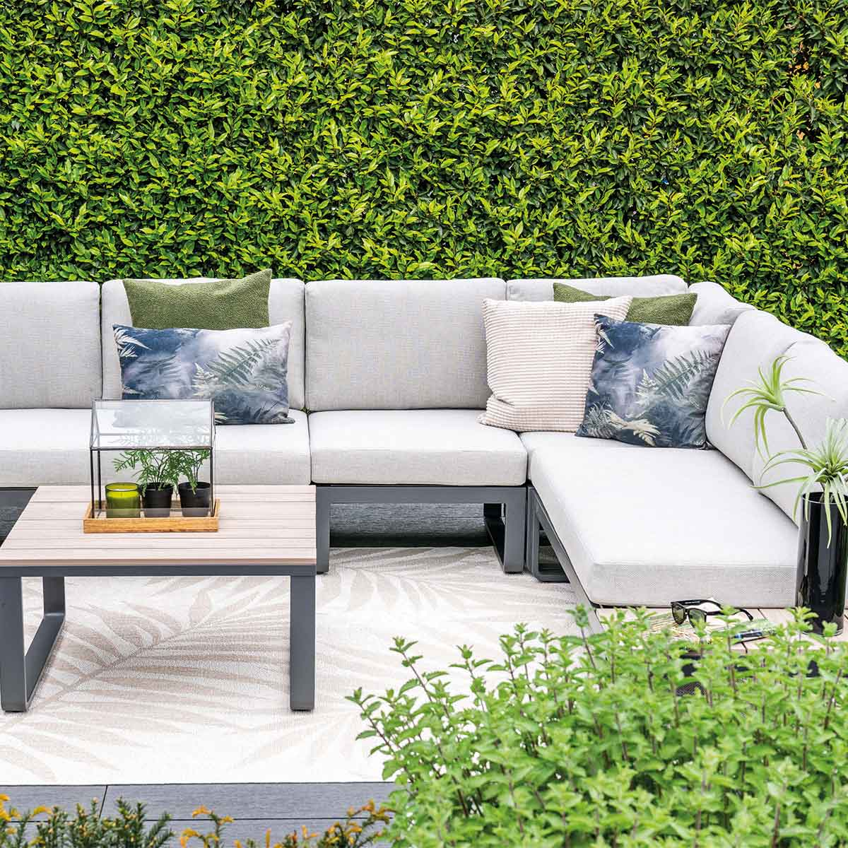 An outdoor seating area with a sofa, pillows, and a low table. There is greenery in the background.