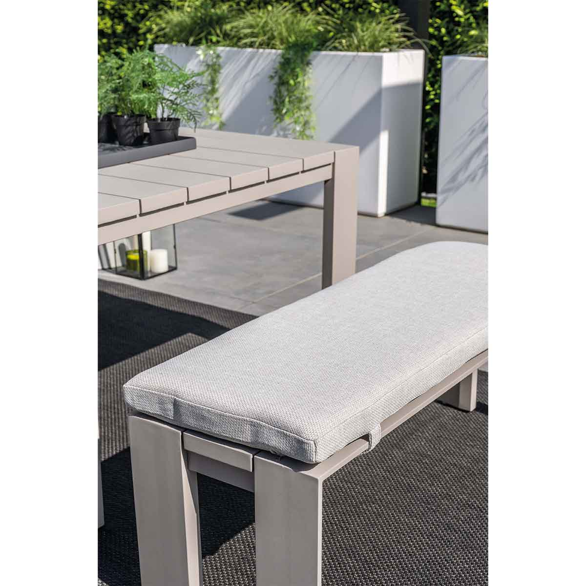 Une table gris clair avec un banc à coussin blanc sur un tapis gris foncé. Conteneurs de plantes blancs en arrière-plan.