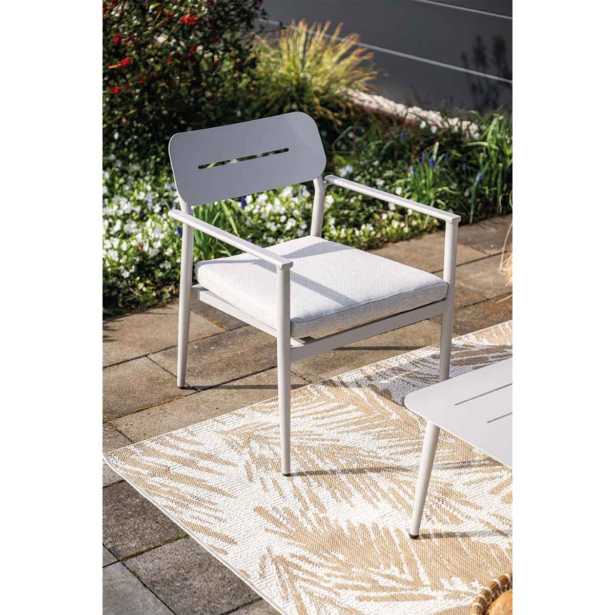 A light gray chair with a cushion sits on a patio. A table and plants are in the background.