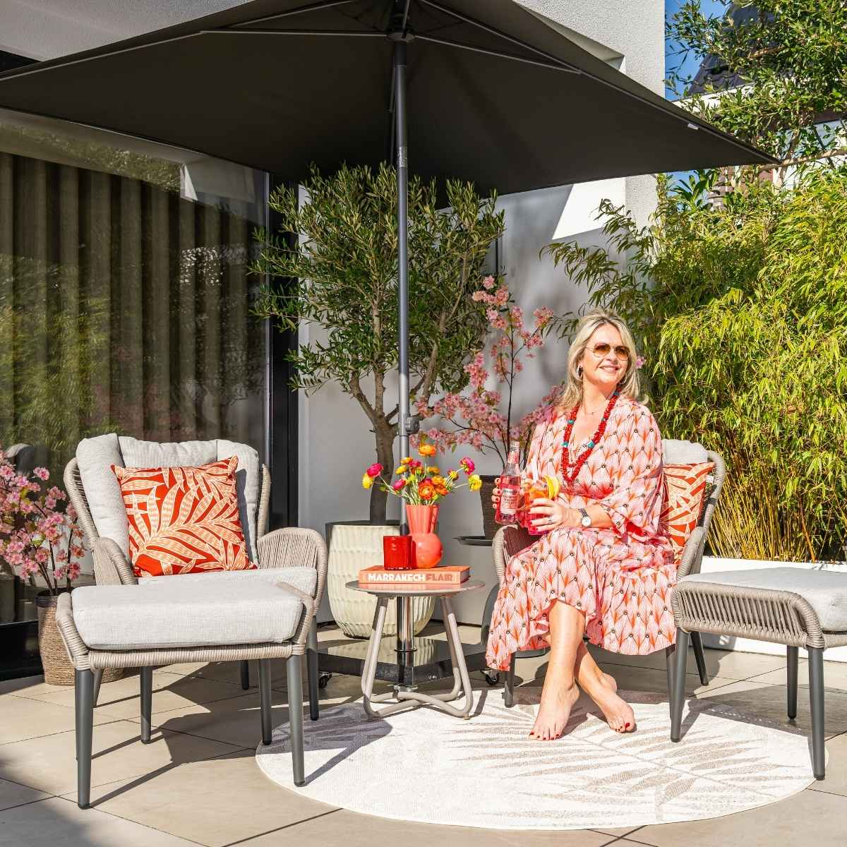 A woman sits under a black umbrella. She's in an outdoor setting with chairs and a table.