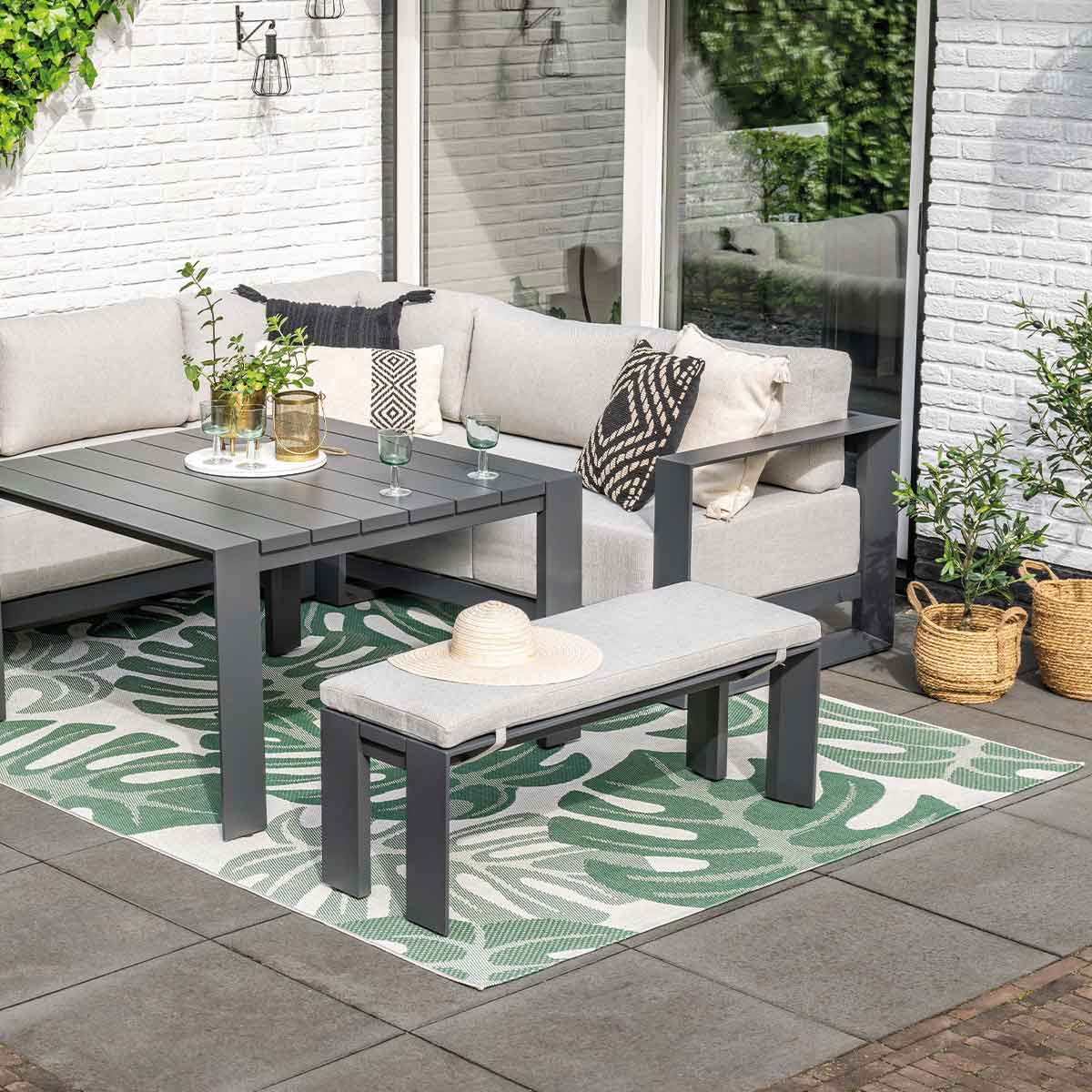 Scène extérieure avec meubles gris, tapis vert et plantes. Canapé, table et banc.