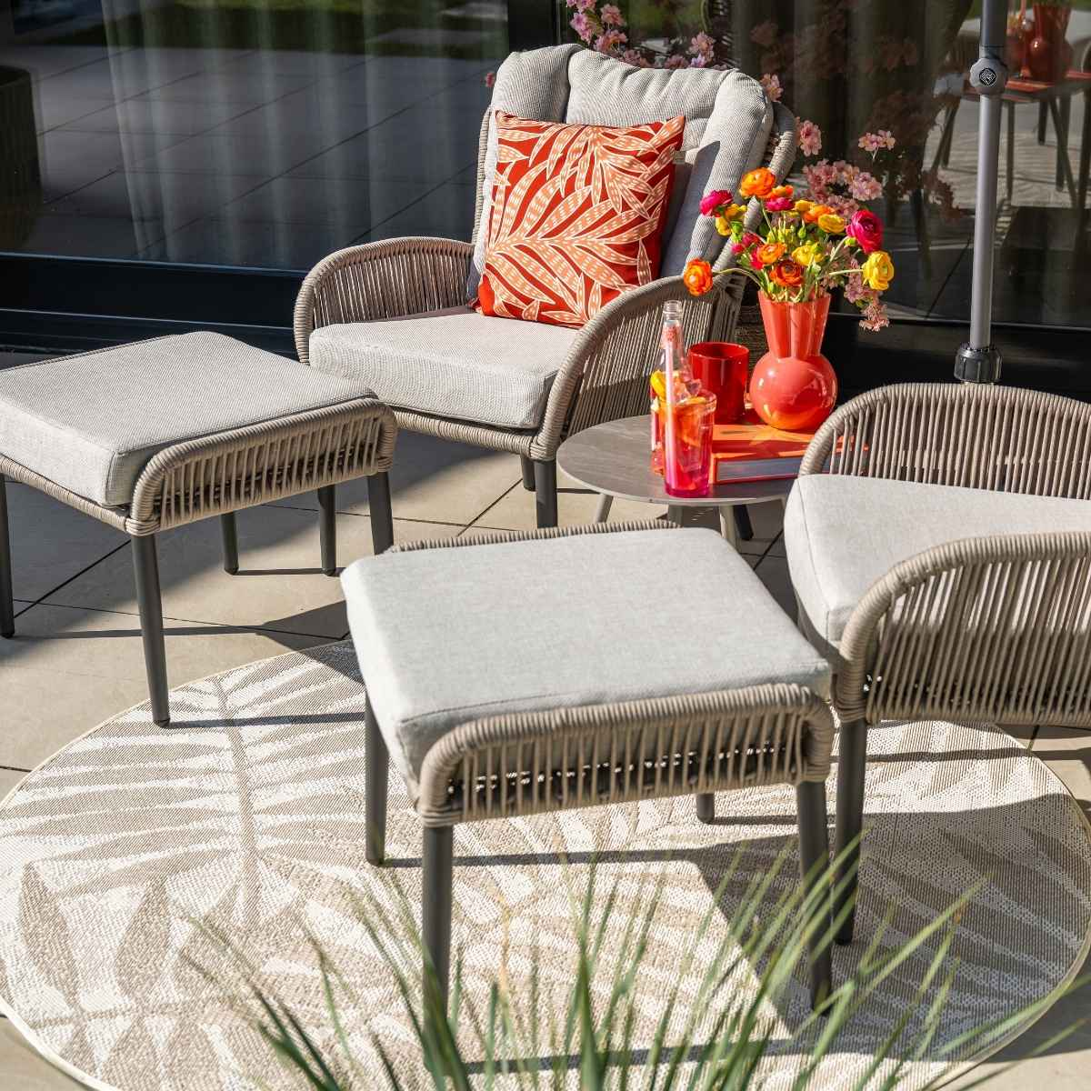 Patio scene with furniture, drinks, and people. Gray chairs, round rug, flowers, and an umbrella.