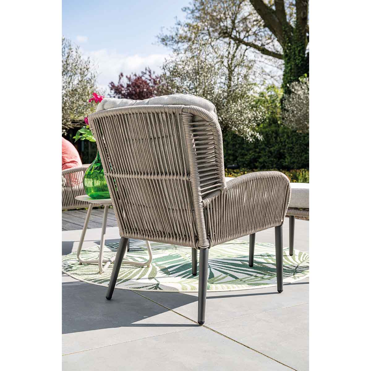 A rattan chair from the back, placed on a patio, with green plants in the background.