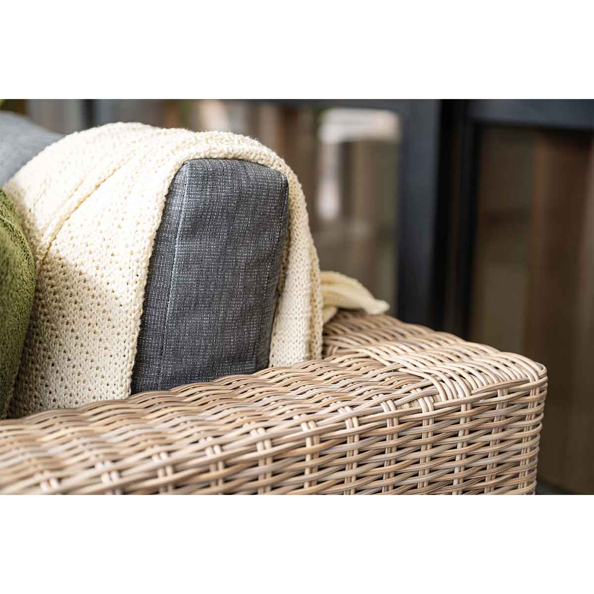 Gros plan sur une chaise tressée et une couverture drapée sur le dossier. Couverture crème sur coussin gris.