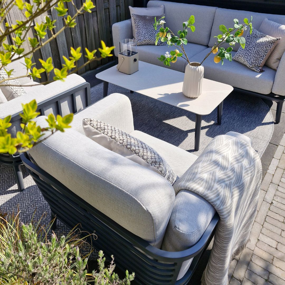 Een loungeset voor buiten. Grijze bank, stoel en tafel. Een deken op de stoel en kussens op de bank.