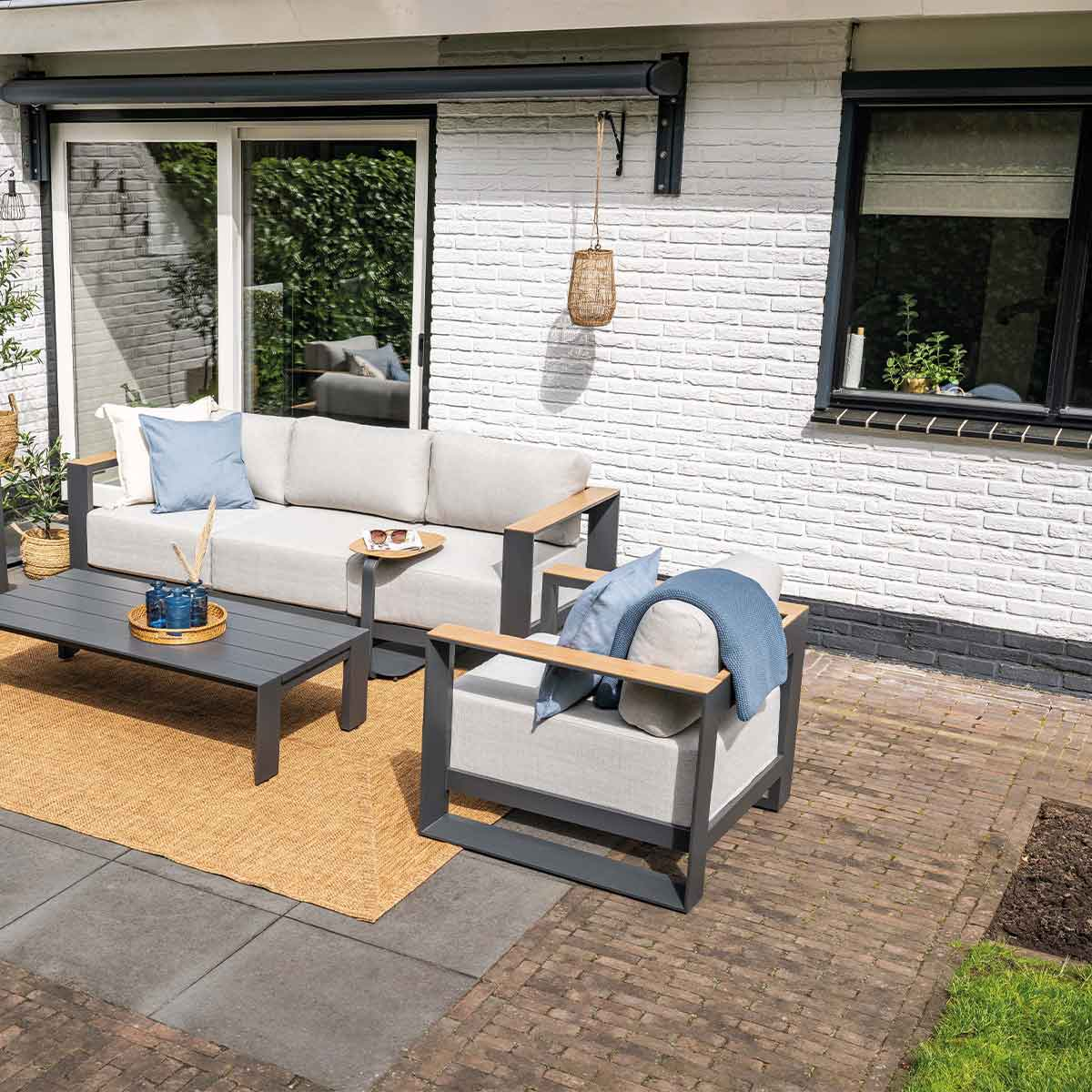 Outdoor patio scene with gray furniture, a rug, and plants. Includes a sofa, chair, and coffee table.