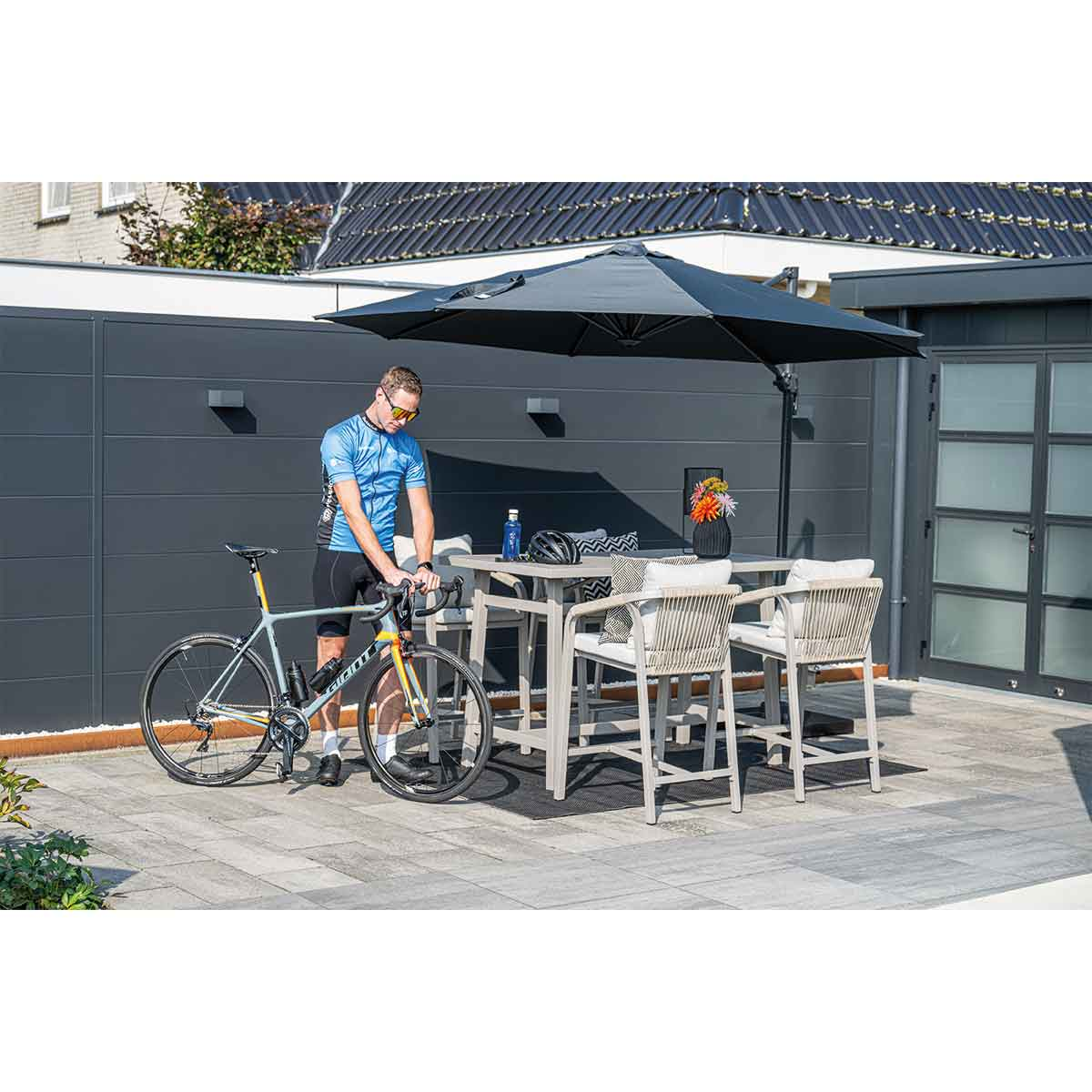 A cyclist next to a table and bar stools, under an umbrella, on a paved patio.