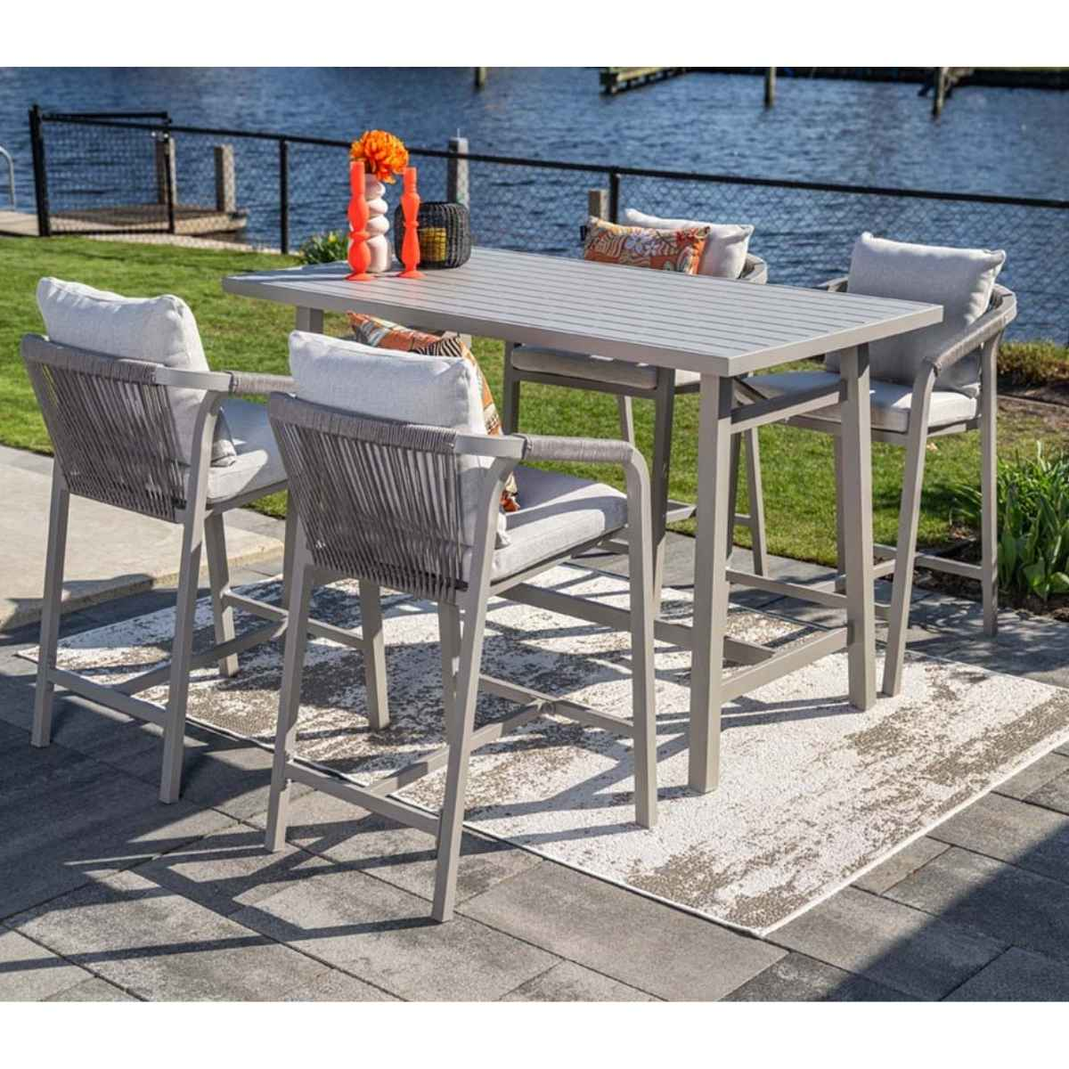 Outdoor dining set with gray high chairs, table, and river view. Includes cushions, rug, and decorative items.