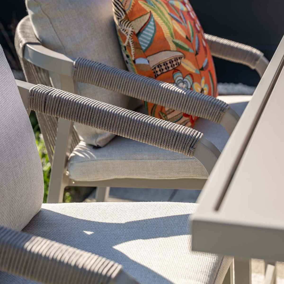Close-up of a gray chair armrest with rope detailing and an orange patterned pillow. Table edge is in view.