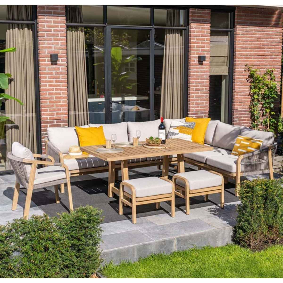 Buitenpatio set. Beige hoekbank, houten tafel, kussens en bakstenen achtergrond.