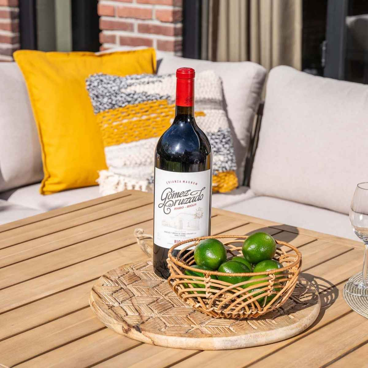 Tafel met wijnfles, limoenen in een mand en kussens op een terras.