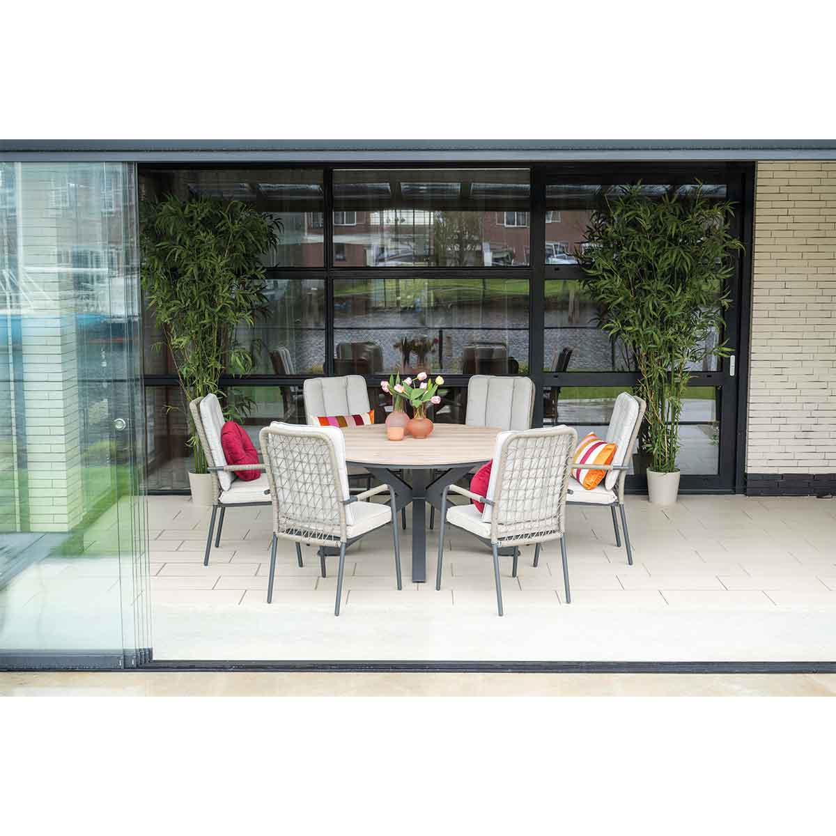 Outdoor dining setting with a round table, six chairs, flowers, and plants, near a glass door.