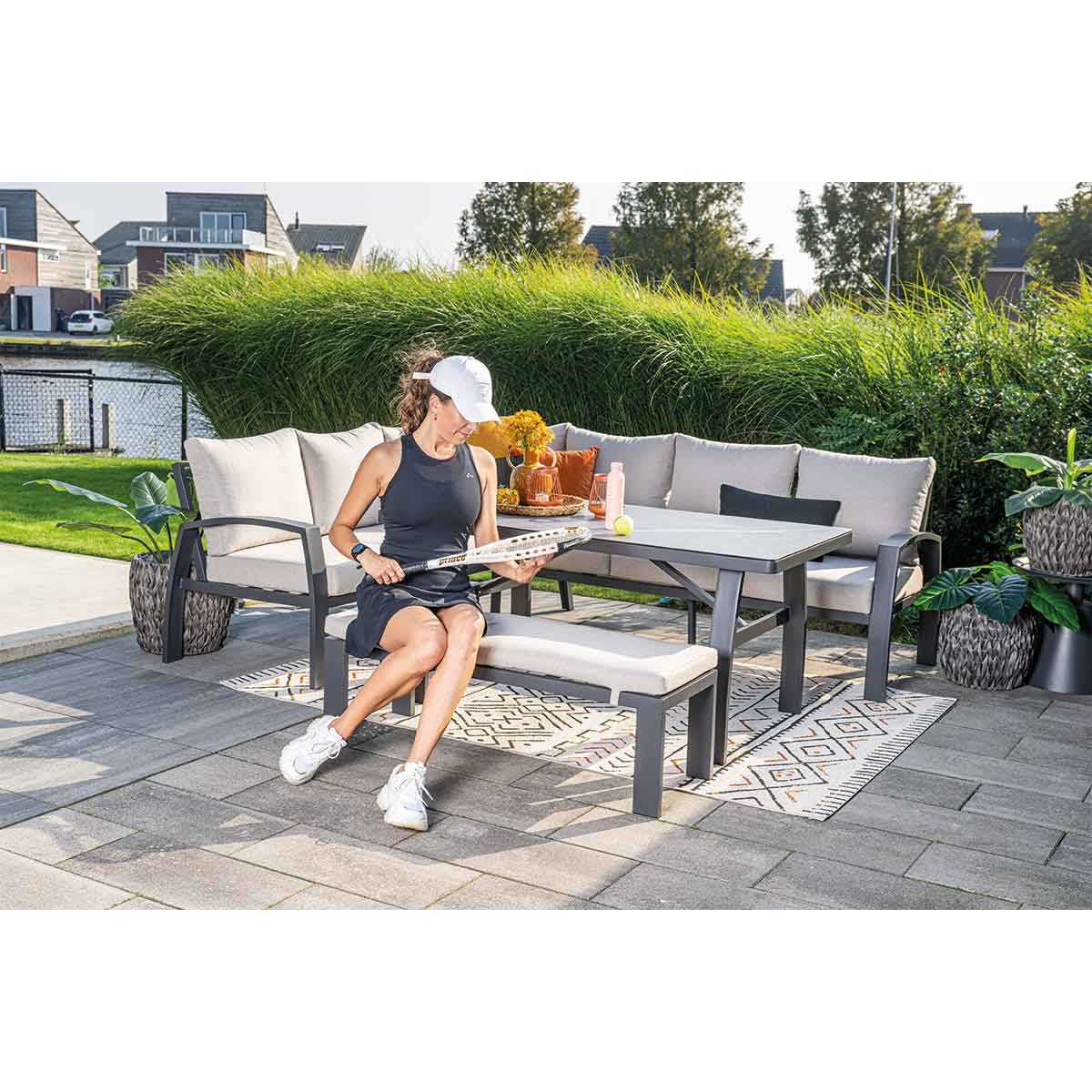 Femme assise sur un ensemble sectionnel extérieur avec une raquette de tennis. Buissons verts et bâtiments en arrière-plan.