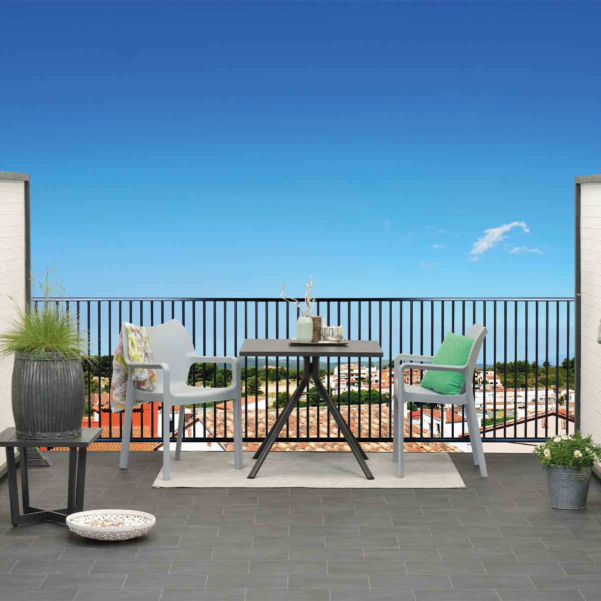 A balcony scene with a table, chairs and a railing. The sky is blue.