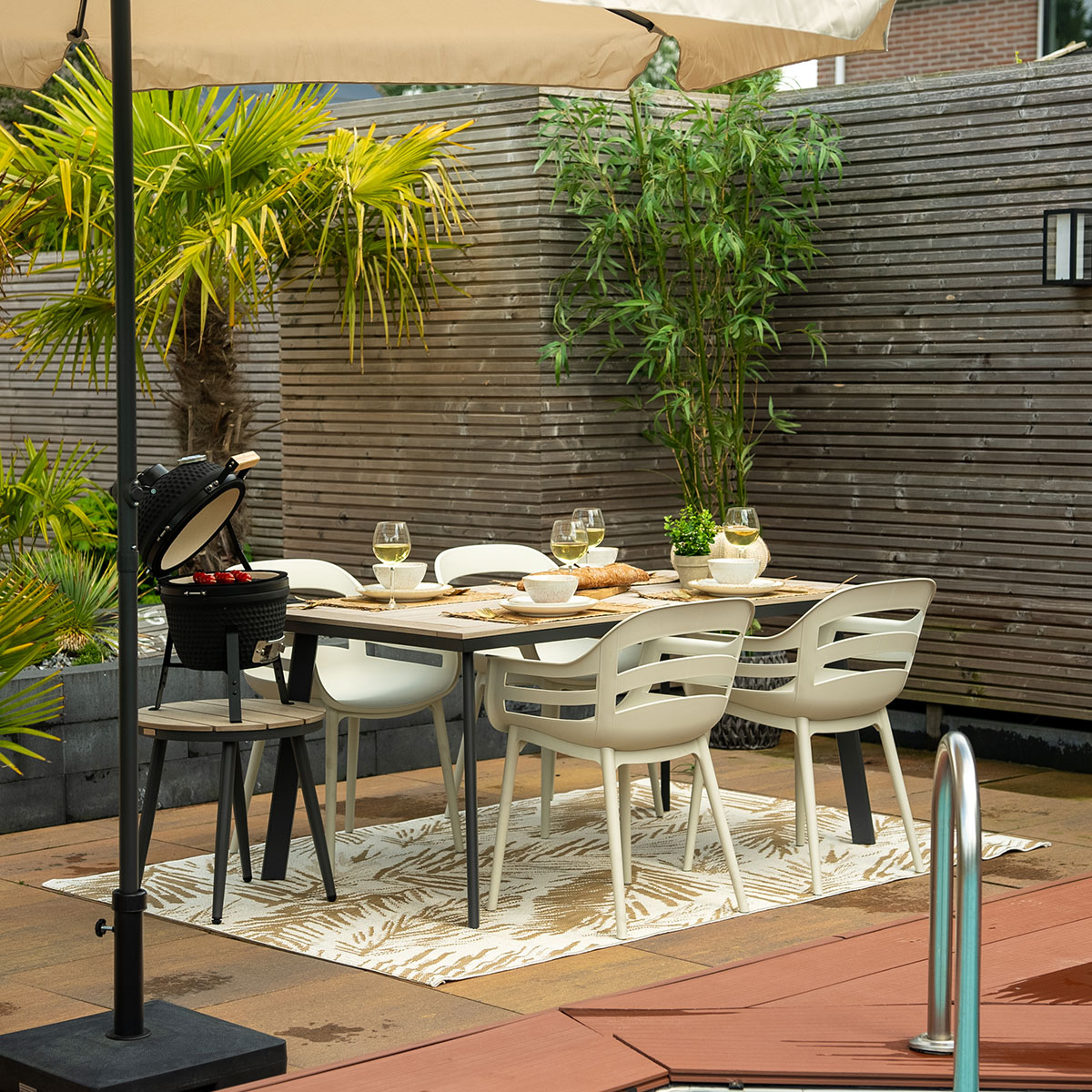 Outdoor dining setting with table, chairs, umbrella, and barbecue. Lush greenery and wood floor.