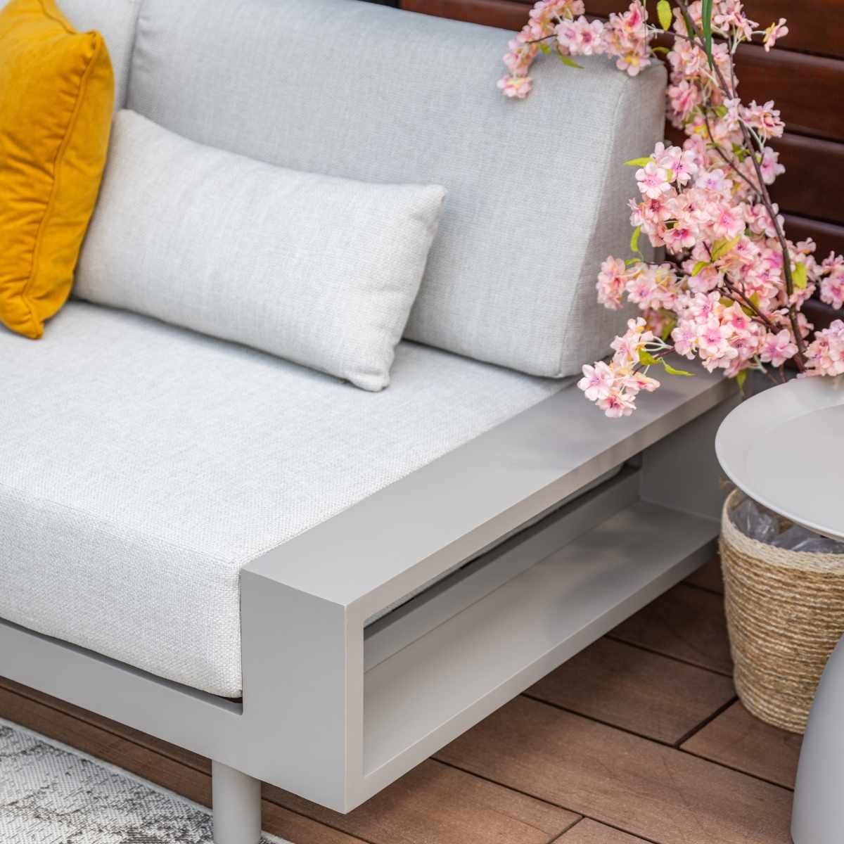 Close-up of sofa with yellow pillow and flowers on side. Wooden deck and white background.