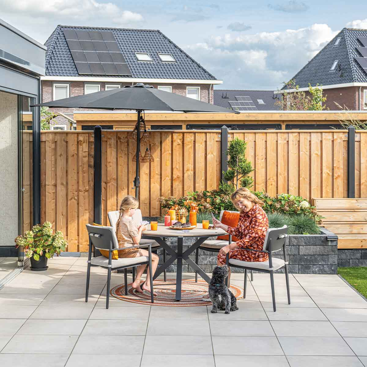 Family playing cards at a table with a dog.  Outdoor setting with houses and an umbrella.
