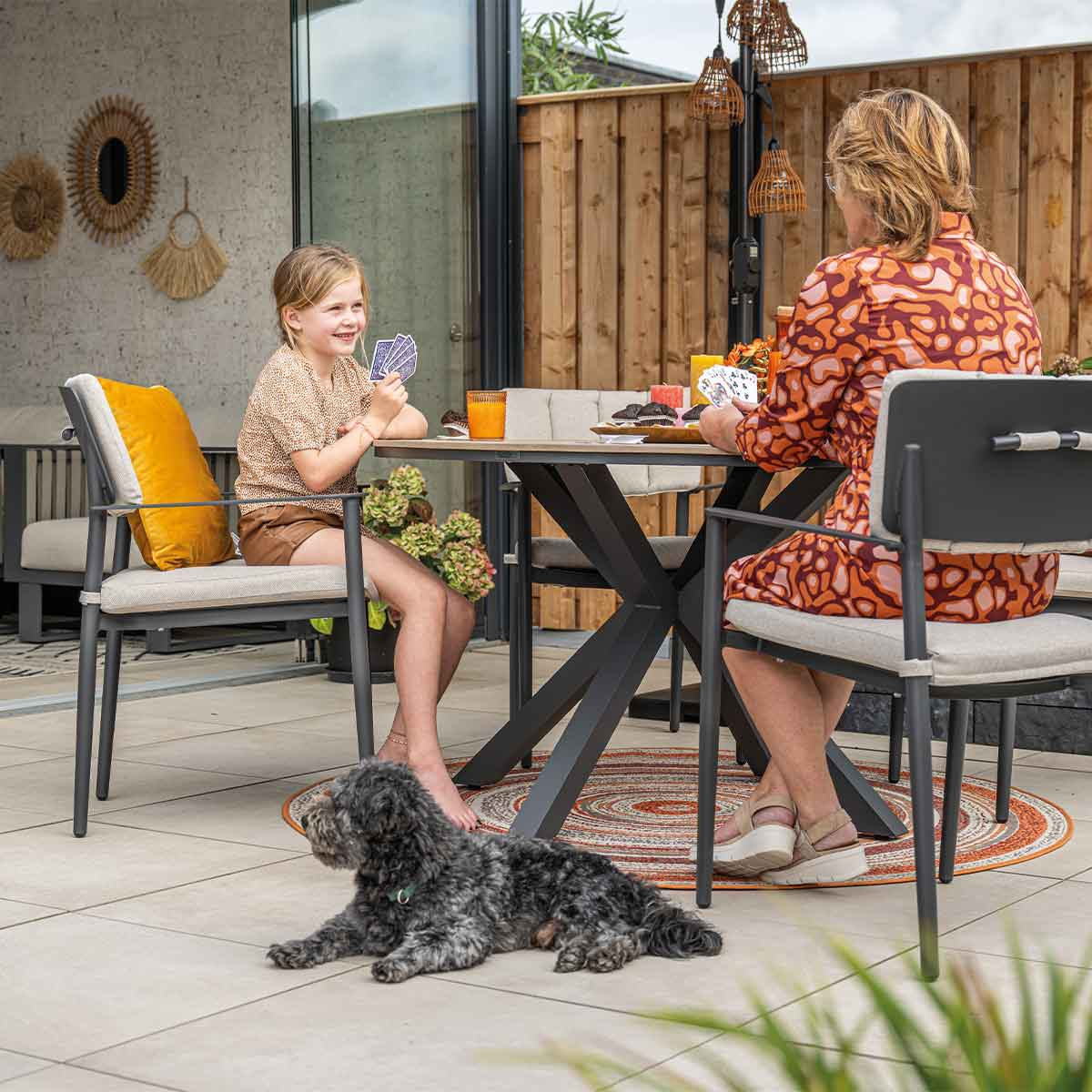 Two people playing cards at a table. Dog lying down. Outdoor setting.