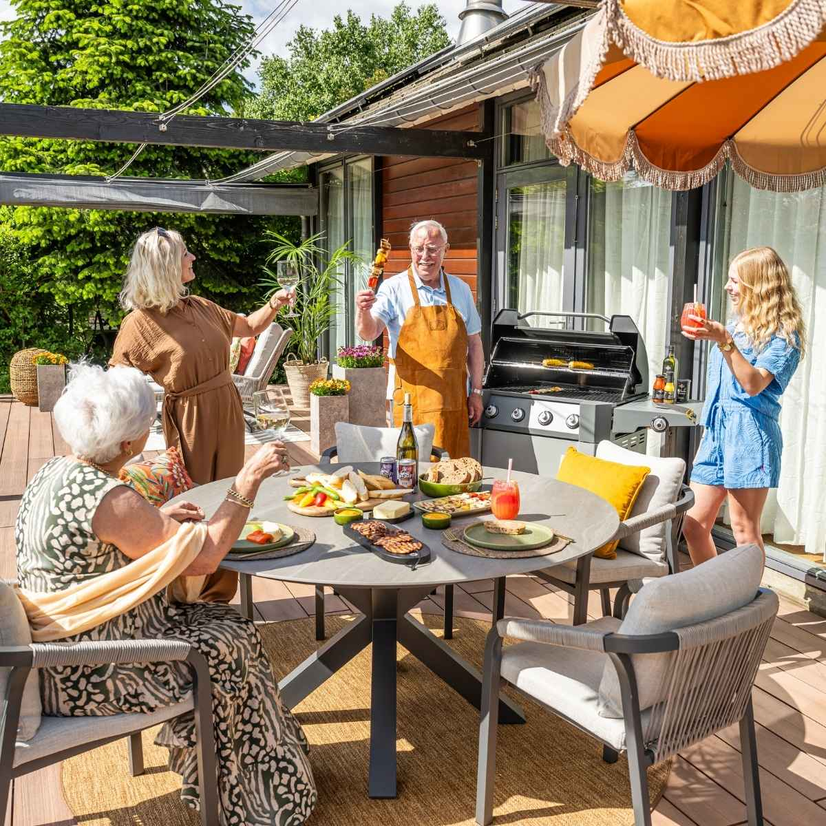 Groep mensen buiten. Ze verzamelen zich rond een tafel met eten en drinken. Grill in de achtergrond.