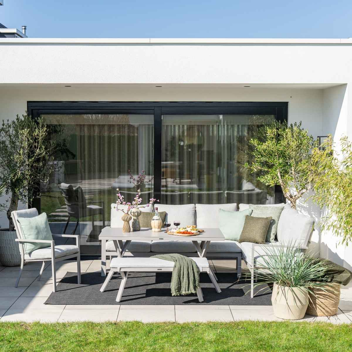 Aménagement de patio avec une table, un canapé, un banc et des plantes, avec une maison blanche en arrière-plan.