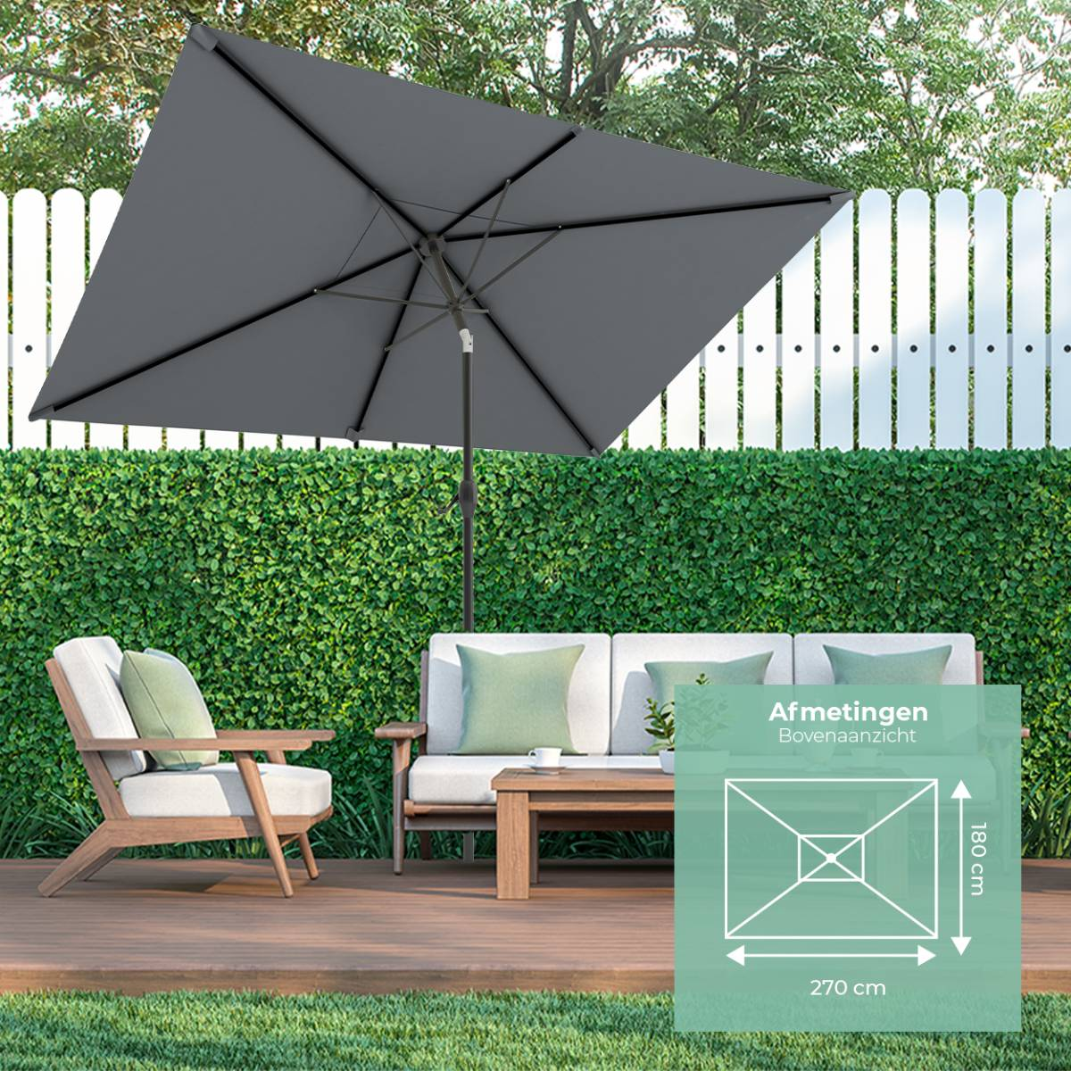 Een grijze parasol boven een houten terras met meubels, groene haag en witte schutting.
