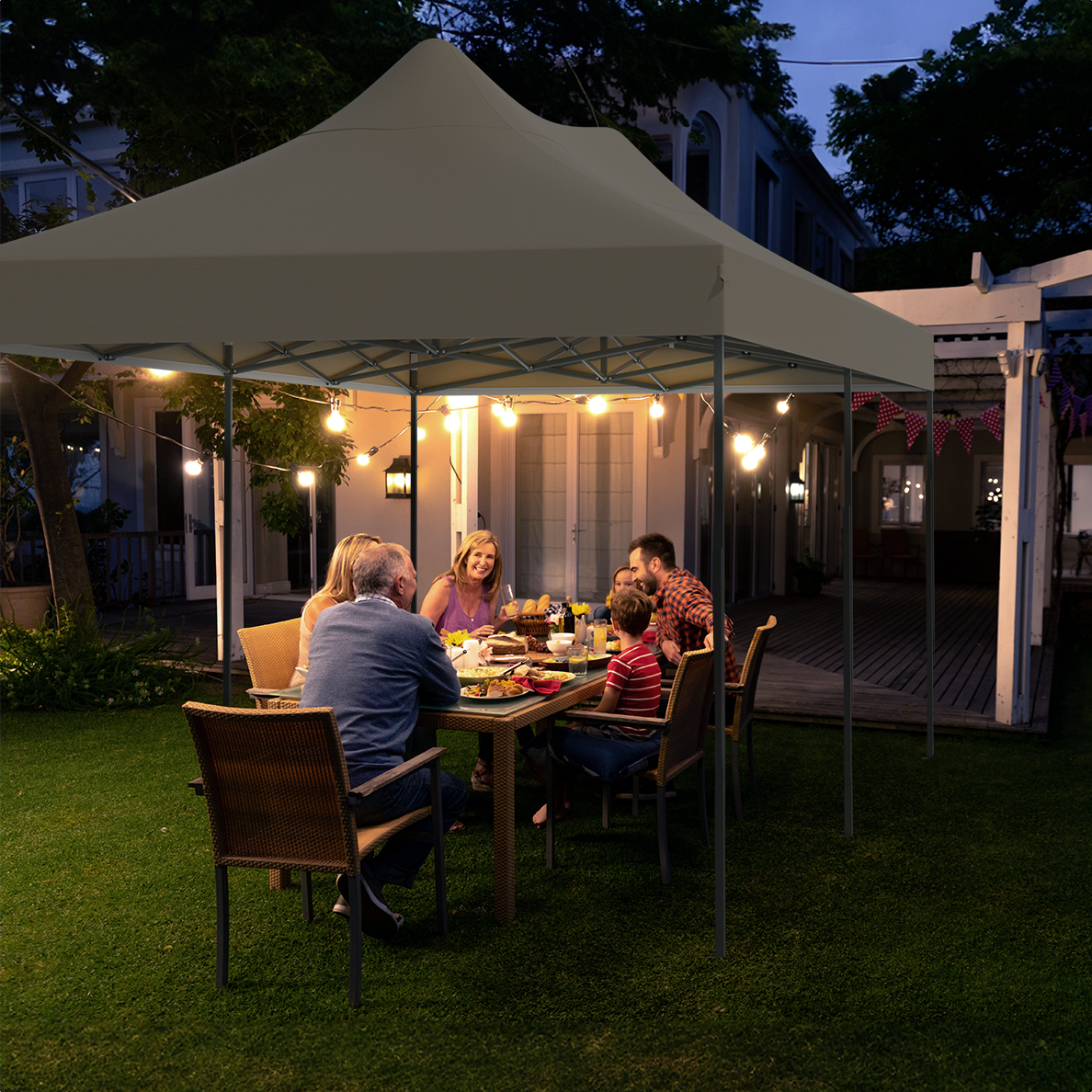 People dining under a tan tent at night, with lights and a house in the background.